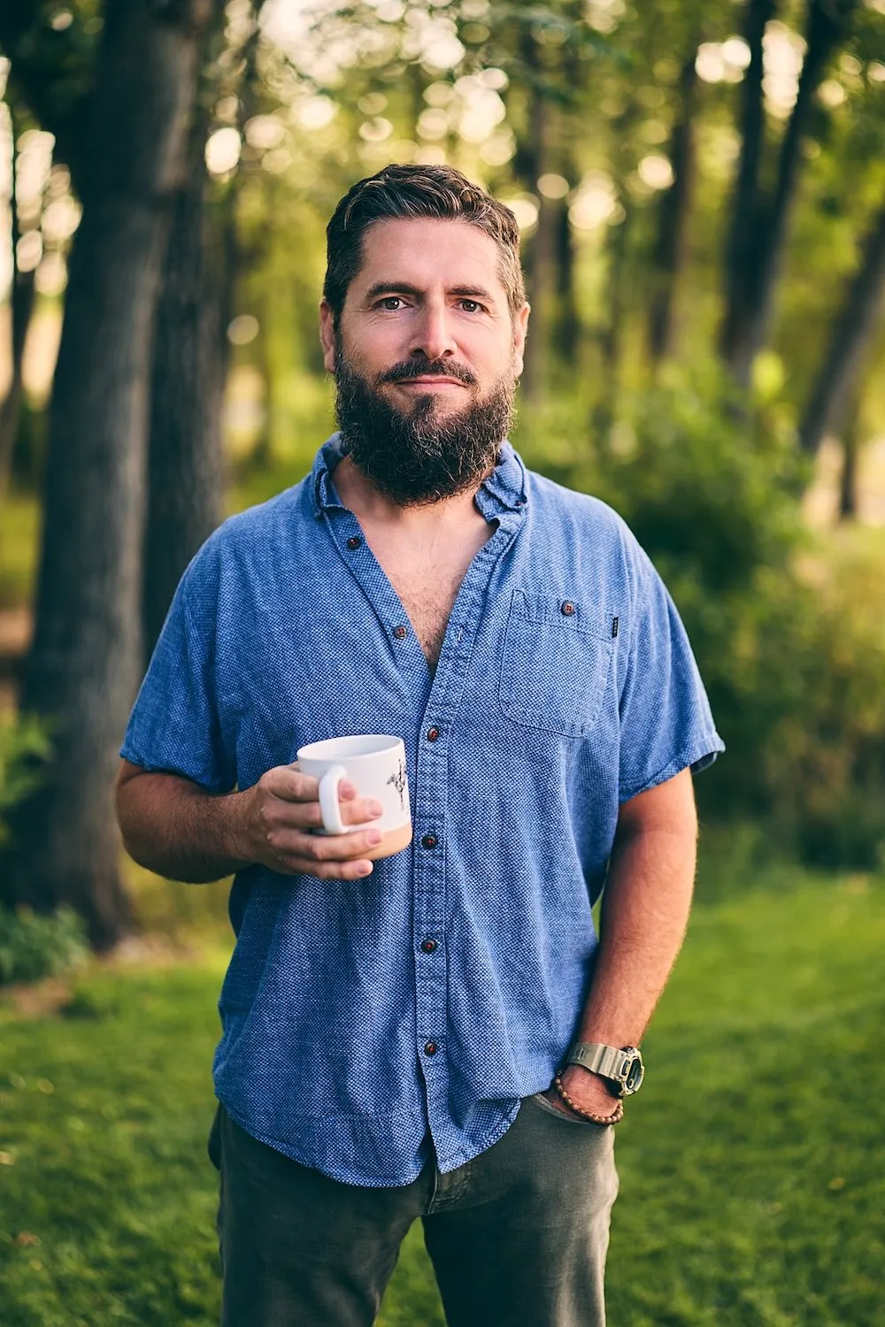 Anthony holding a coffee mug in a sunlit grove of trees
