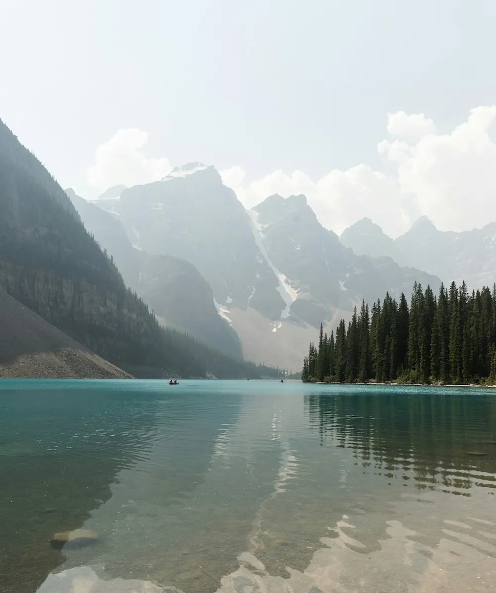 A turquoise mountain lake with pine trees and hazy peaks